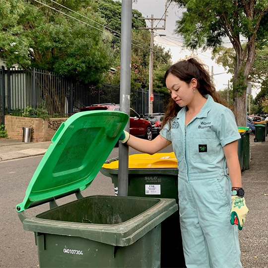 Person dressed in a light teal work uniform inspecting a green FOGO bin